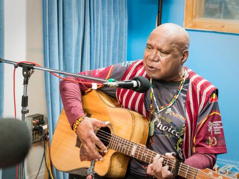 Indigenous singer/songwriter Archie Roach, pictured seated with a guitar in front of a microphone stand