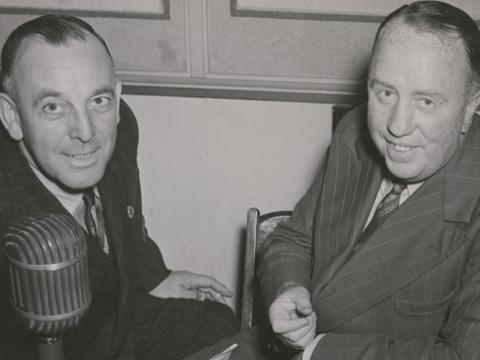 Two men sit next to each other looking at the camera. There is a microphone in front of them.