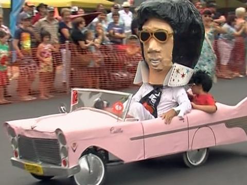 Pink car being driven in a parade by someone in an Elvis Presley costume with a giant Elvis head.