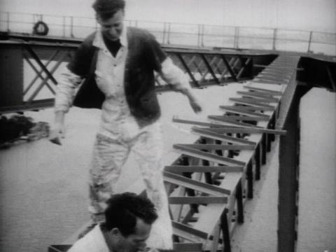 Black and white still of one bridge rigger preparing to step over another rigger who is sitting down on a steel truss of the Sydney Harbour Bridge. They're not wearing any kind of safety harnesses and there are no visible safety precautions in place.