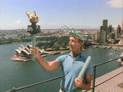 Paul Hogan dressed as the Statue of Liberty stands on top of the Sydney Harbour Bridge with the Opera House and Circular Quay in the background