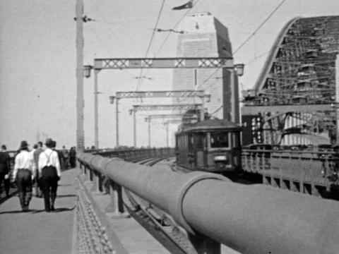 Black and white still of people walking across the newly-opened Sydney Harbour Bridge in 1932. A tram rolls past on the tracks across the barrier from pedestrians.