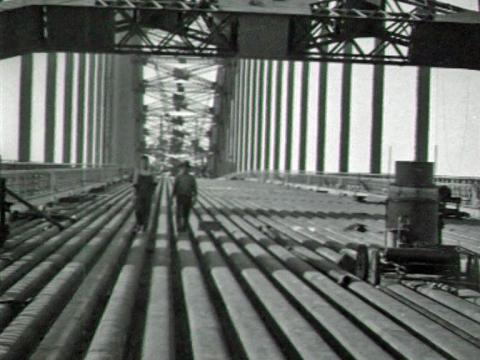 Black and white image of two men walking along the main arch decking and troughing of the Sydney Harbour Bridge before it has been concreted and asphalted.