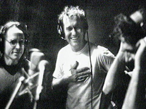 Phil Small, Jimmy Barnes and Ian Moss from the band Cold Chisel standing in a recording studio in front of microphones and all wearing headphones. 