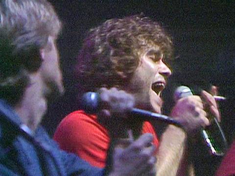 Close up of Jimmy Barnes in 1980 singing into a microphone. In the foreground is singer Mick Pealing who is also singing and has his head turned away from the camera. 