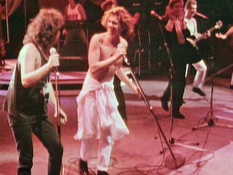 Jimmy Barnes and Michael Hutchence on stage at nighttime with lights at the back, Jimmy is dressed in black jeans and singlet and Michael is wearing white pants and no shirt. Both are singing into microphones.