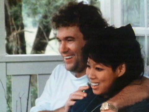Jimmy Barnes, seated with his arm around his wife Jane. Both are smiling and facing to the left of screen. They appear to be seated outside on a front deck of a house. 
