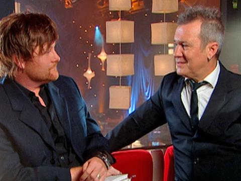 Music television host Chit Chat talks to singer Jimmy Barnes. The two men are both wearing black suits and facing each other. 