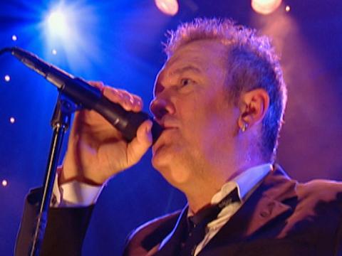 Head and shoulders shot of Jimmy Barnes singing into a microphone, he is wearing a suit, white shirt and black tie with blue and purple lighting in the background.