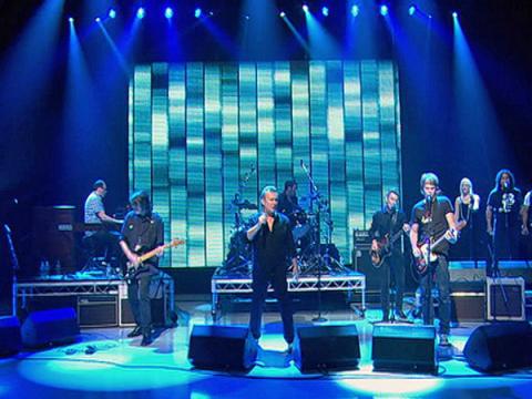 Wide shot of Jimmy Barnes (centre) on a TV studio stage surrounded by band members. The TV studio is flooded in different shades of blue lights. Barnes is dressed all in black and holding a microphone.