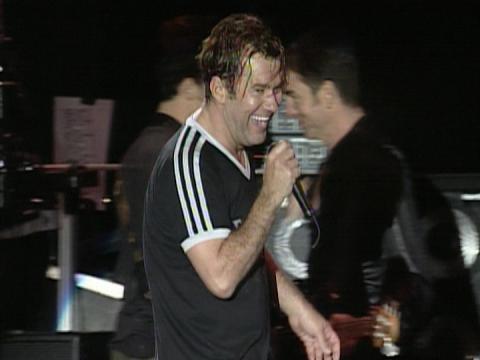 Jimmy Barnes pictured from the waist up on stage wearing a black t-shirt with white stripes on the sleeves, holding a microphone and smiling.