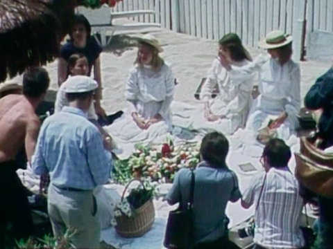 A group of people, including some young women in early 20th century period costume seated in a circle having a picnic.