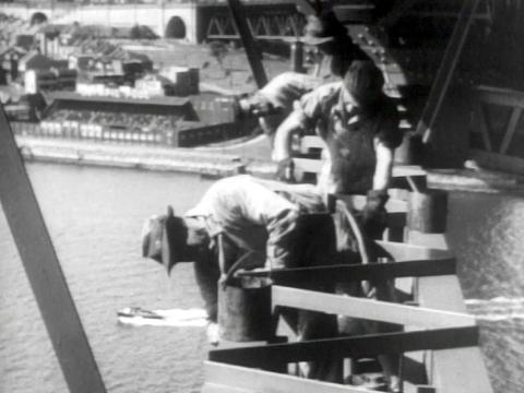 Four men paint the Sydney Harbour Bridge. You can see a boat passing in the harbour below. They are all wearing hats.