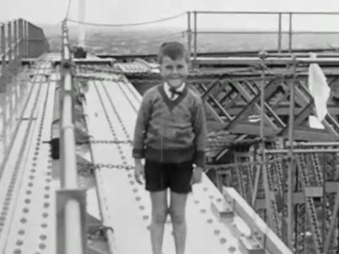 Eight year-old Kenneth Jones stands high above the ground on top of the arch of the Sydney Harbour Bridge smiling. He's wearing a bow tie and school jumper and shorts. There is no evidence of harness or safety precautions for the young lad.