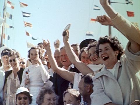 A crowd of people wave enthusiastically at Queen Elizabeth II on her visit to Hobart in 1954. There are flags in the background.