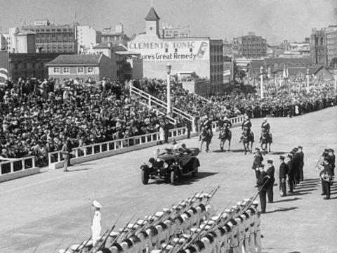 Wide shot of the Premier of NSW's car arriving at the opening of the bridge, men on horseback behind, navy men standing to attention, crowd in the stands