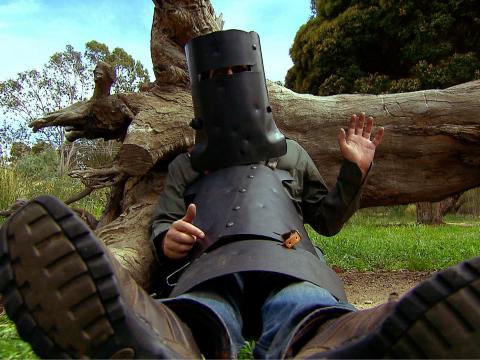 An actor dressed in Ned Kelly's armour lying against a tree trunk.