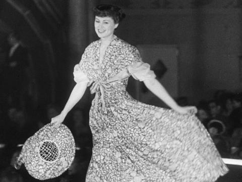 A model is wearing a floral dress and holding a matching hat as she walks.