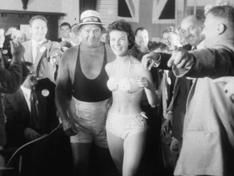 A woman in a bikini is being led out of a room of men in suits by a lifeguard.