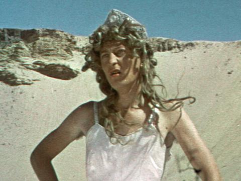Graham Kennedy in a long blonde wig and dress standing in the desert
