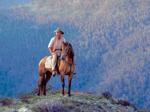 Jim Craig (played by Tom Burlinson) sits in the saddle on a horse on the top of a hill with rugged mountains in the background, in a scene from 'The Man From Snowy River'