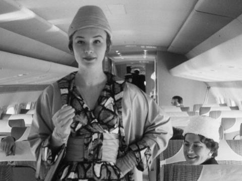 A model wearing a hat parades down an aisle in an aircraft. A passenger turns her head to look.