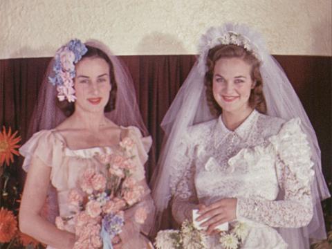 Two women are standing next to each other looking at the camera. One is wearing a bridal gown and veil, the other is a bridesmaid.