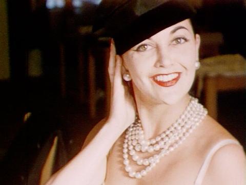 A woman wearing pearls, a hat and not much else smiles at the camera.