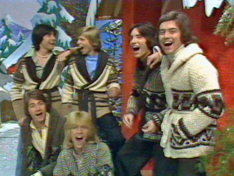 A group of six young men in a studio singing. The studio is decorated with a snowy, winter theme.