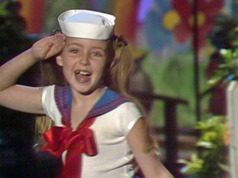 A young girl performing a song in a cute sailors outfit salutes as she sings to camera. 