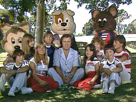 A man sits in a park surrounded by a group of teenagers and some animal mascots. They are all smiling at the camera.
