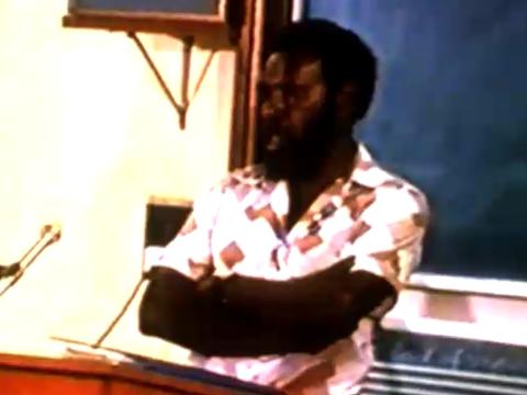 Eddie Mabo with arms crossed speaking in front of a blackboard.