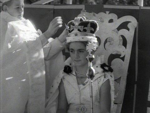 A schoolgirl sitting in an ornate chair dressed as The Queen while a boy places a crown on her head.