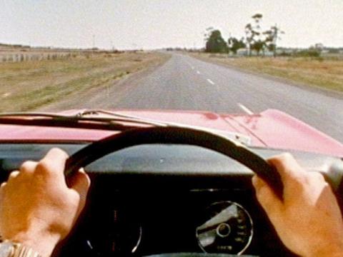 Point of view of a man driving a vintage XT Falcon on a straight stretch of road. We see his hands on the steering wheel.