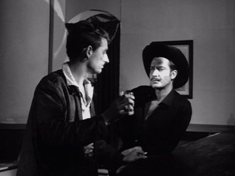 A man in a black western outfit, complete with hat is talking to another man who's drinking at a bar.