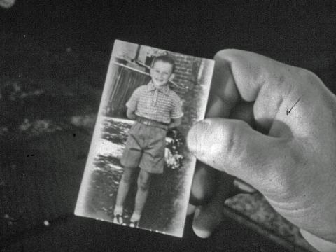 A close up of a man's hand holding a small photograph of a young boy. The boy in the photograph is wearing a shirt and shorts and is smiling directly at the camera. 