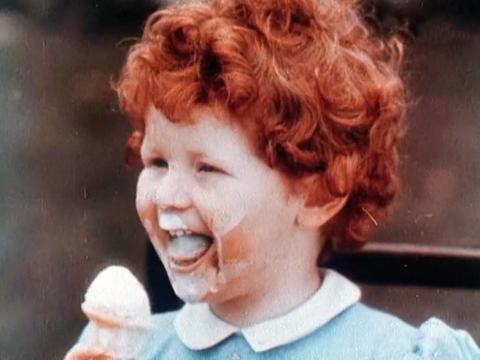 Close up of a young child with curly red hair eating an ice cream. She has the ice cream all over her face and is smiling.