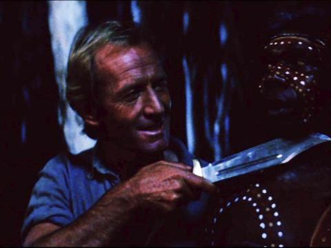 Video still from Crocodile Dundee showing Mick Dundee (Paul Hogan) holding a knife jokingly to Neville's (David Gulpilil's) throat