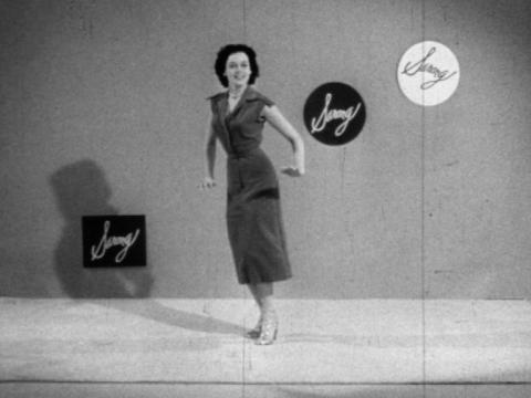 A black and white still of a woman dancing wearing a dress.