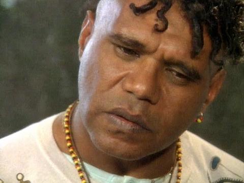 Close up of Indigenous singer-songwriter Archie Roach. He's wearing a beaded necklace and white t-shirt, his head is slightly tilted with his eyes looking downward.