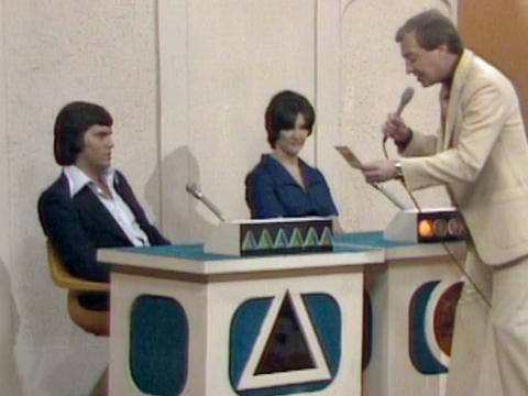 Game show set in TV studio with two contestants and a compere holding a microphone.