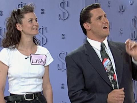 A young woman stands next to a game show host. They are both looking up and to their left at something off camera.