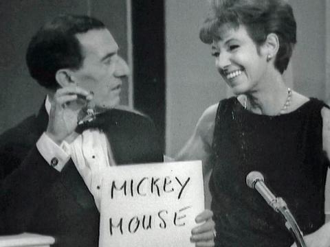 A middle-aged husband and wife sit looking at each other and smiling. He is holding a sign that says Mickey Mouse  