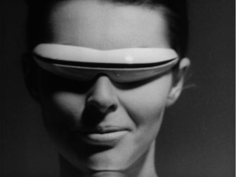 A woman wearing futuristic glasses from the 1960s which are like a band around her eyes. She is smiling knowingly.