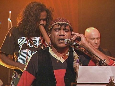 Archie Roach pictured in front of a microphone on stage. Behind him is another musician playing didgeridoo. 