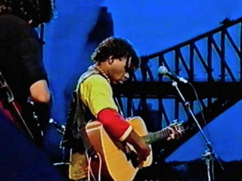 Archie Roach pictured from the side holding a guitar and standing in front of a microphone. He's wearing a yellow shirt and black vest. The Sydney Harbour Bridge can be seen in the background. 
