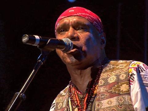 Archie Roach pictured from the chest up in front of a microphone, wearing a red bandanna on his head and a patterned vest.