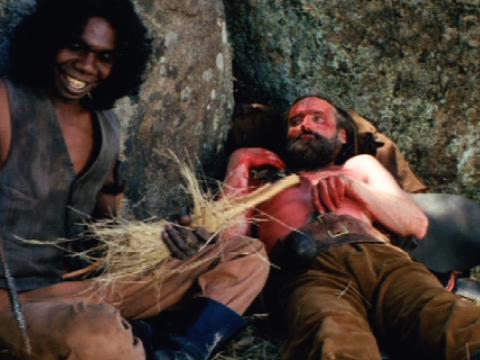 A man is lying on his back looking at another man who is smiling. The smiling man is Aboriginal and is holding a spear. The other man is white and is covered in blood.