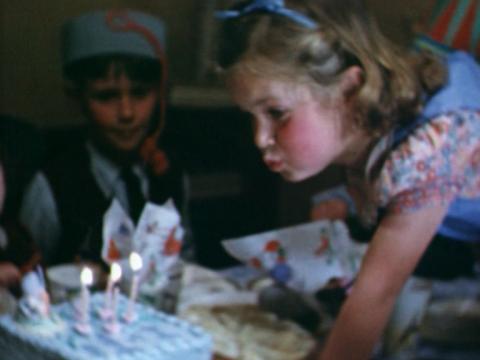 A little girl wearing a blue party dress and matching headband blowing out the birthday candles on her cake