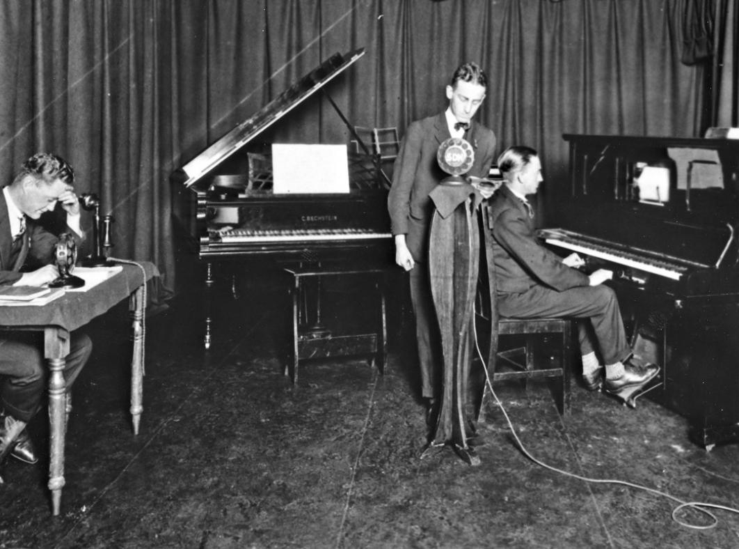 Recording a radio program from a home recording studio in 1927. One man sits at a desk, another at a piano while a third stands behind a microphone.
